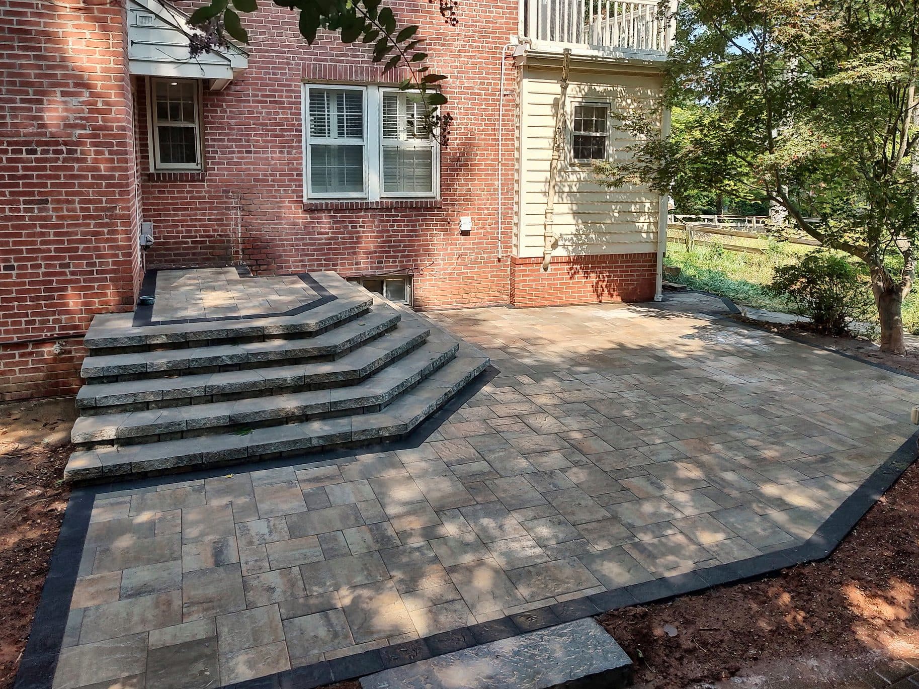 Angled view of a paver patio and wraparound step landing with a charcoal inlay border against a brick home.