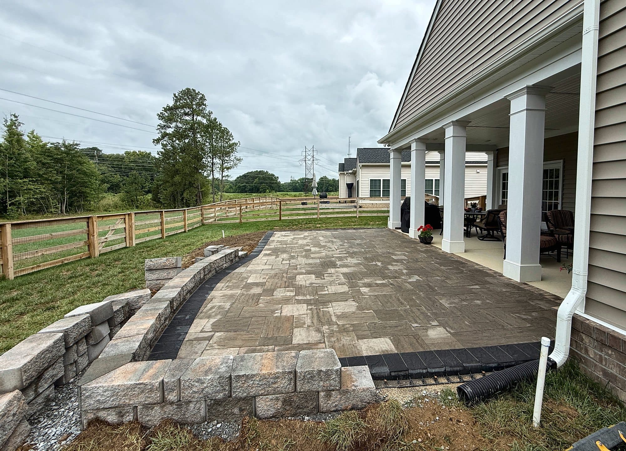 Segmental wall and steps under construction beside a new paver patio, with drain pipe being routed from a downspout.