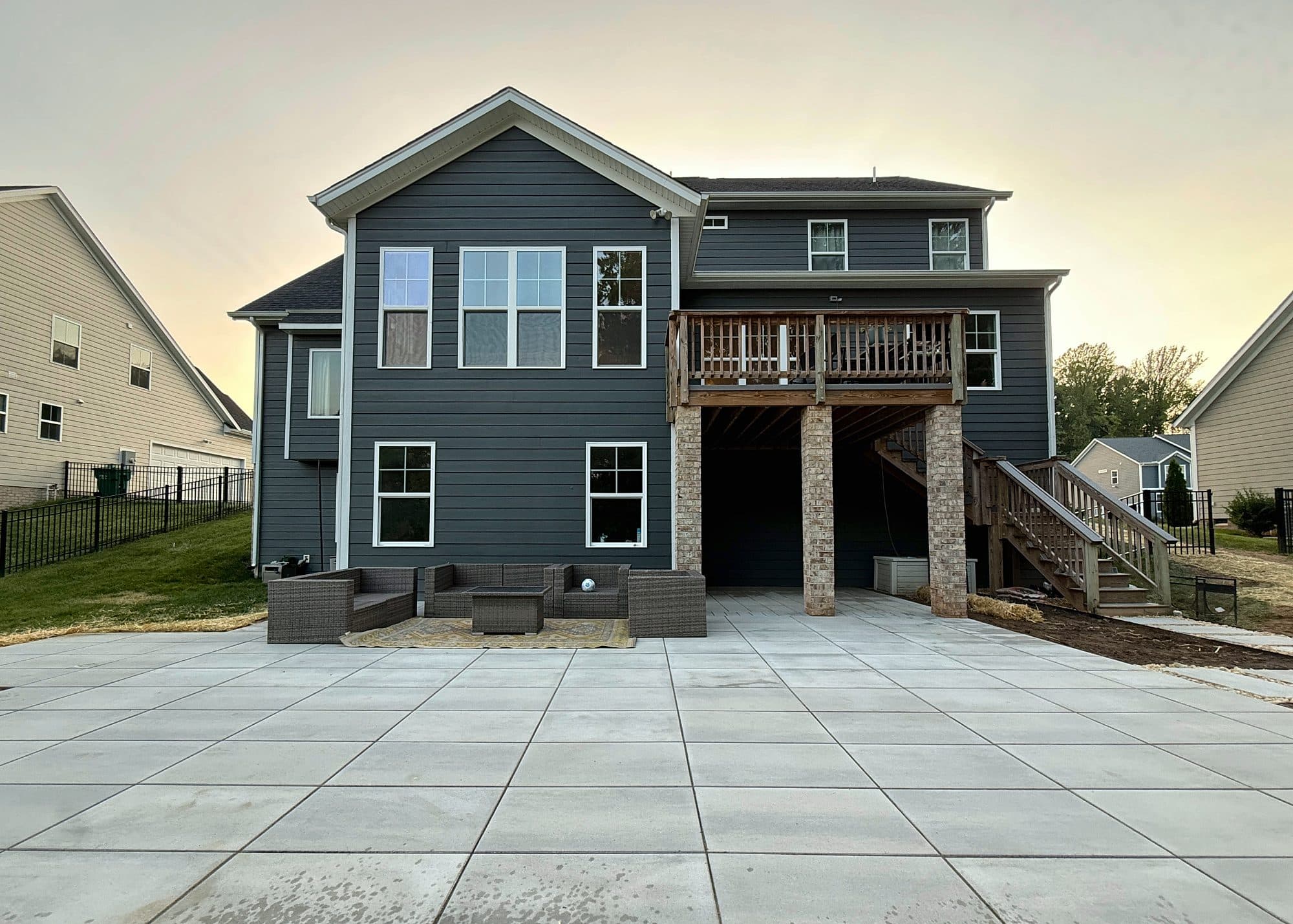 Large-format slab patio laid in a clean grid behind a two-story home at dusk, with a seating area beneath the deck.