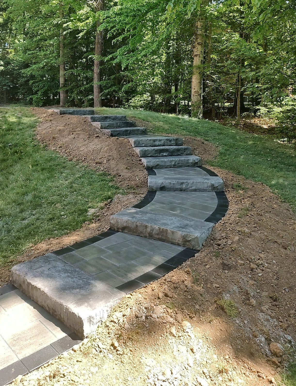 Stone slab steps curving up a wooded slope, with large-format paver landings edged in a charcoal border.
