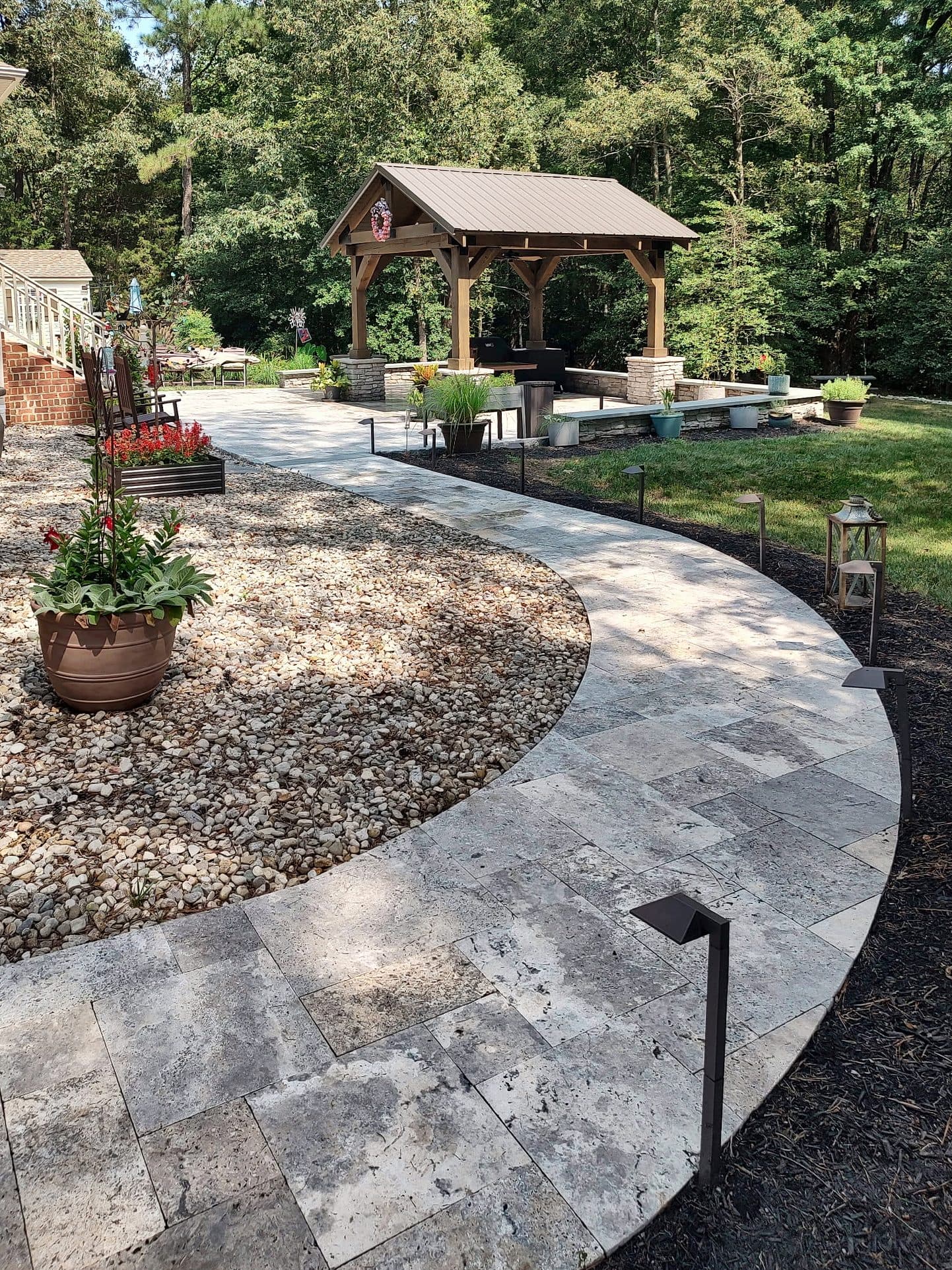 Curved travertine walkway lined with low-voltage path lights, leading to a timber-frame pavilion with stacked-stone column bases.